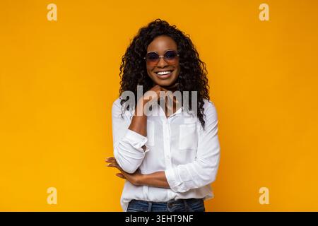 Femme souriante portant des lunettes de soleil pose en toute confiance devant un fond jaune vif Banque D'Images