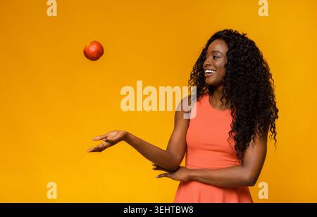 Femme en robe rose équilibre pomme rouge contre fond jaune vibrant dans un moment ludique Banque D'Images