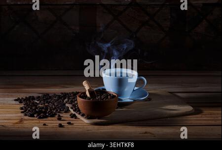 Moody Still Life de café chaud fraîchement infusé dans une tasse en céramique blanche avec de la vapeur visible, placée à côté d'un bol en bois de grains de café torréfiés. Banque D'Images