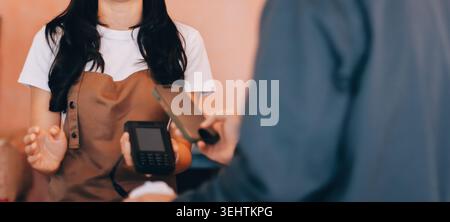 Barista adulte senior asiatique recevant un paiement mobile de la part d'un client utilisant un smartphone dans un café démontrant une transaction sans espèces moderne dans Cozy lo Banque D'Images
