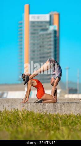 Deux jeunes acrobates réalisant un impressionnant équilibre acroyoga posent à l’extérieur sur une structure en béton avec un bâtiment urbain et un ciel bleu clair à l’arrière Banque D'Images