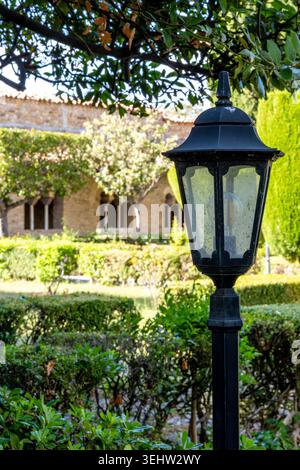 Vieste - Pouilles - Italie - 29-08-2025 : lanterne de jardin en métal noir se dresse sous des branches feuillues, avec des haies taillées et un bâtiment en pierre avec des fenêtres cintrées légèrement derrière. Banque D'Images