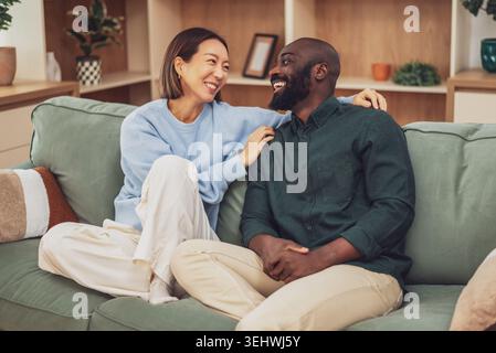 Un joyeux couple multiracial est assis étroitement ensemble sur un canapé vert, riant et embrassant dans un salon confortable et moderne. Banque D'Images