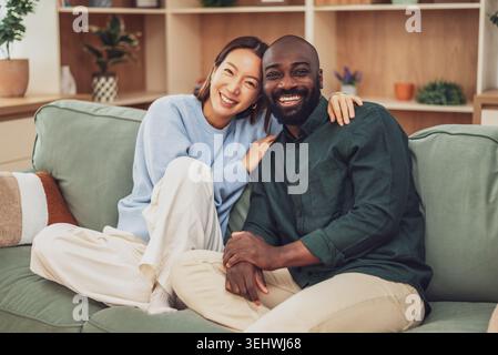 Un joyeux couple multiracial est assis étroitement ensemble sur un canapé vert, riant et embrassant dans un salon confortable et moderne. Banque D'Images