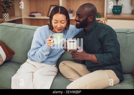 Couple interracial heureux partage un moment joyeux tout en se relaxant à la maison. Ils boivent du café et parlent. Banque D'Images