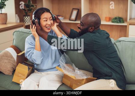 Un couple souriant est assis ensemble sur un canapé vert, ouvrant des boîtes de livraison tandis que l'homme tient un casque sans fil partageant un moment joyeux pendant que le magasinage Banque D'Images