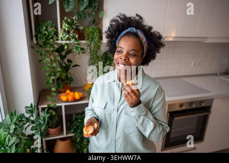 Heureuse jeune femme afro-américaine souriante regardant la fenêtre tenant la mandarine profiter du week-end à la maison Banque D'Images