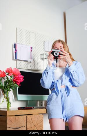Femme blonde souriante utilisant un appareil photo vintage à l'intérieur, scène de style de vie féminine moderne avec espace de travail à la maison et pivoines lumineuses. Banque D'Images