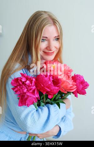 Belle femme blonde avec bouquet de pivoines posant dans une tenue pastel, portrait de style de vie féminin avec fond blanc propre. Banque D'Images