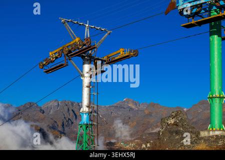 Une tour de téléphérique mécanique avec des roues de poulie et des câbles en acier posée contre une chaîne de montagnes accidentée et des nuages bas sous un ciel bleu clair. Banque D'Images