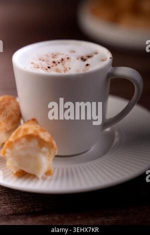 gros plan d'une tasse de cappuccino blanc fumant avec de la mousse mousseuse et des taches de cacao sur une soucoupe striée, accompagnée de bouffées à la crème crémeuses lors d'une guerre Banque D'Images