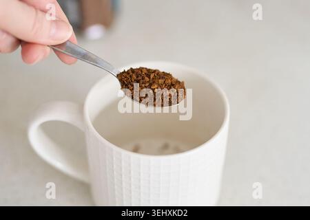 Cuillerée de granulés de café instantané tenue sur une tasse blanche vide sur le comptoir de cuisine avec pot flou en arrière-plan dans le cadre de préparation en gros plan à la lumière du jour. Banque D'Images