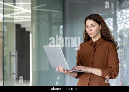 Femme d'affaires professionnelle debout dans un cadre de bureau contemporain, axée sur la saisie sur son ordinateur portable, représentant la flexibilité de travail moderne et l'intégration de la technologie Banque D'Images