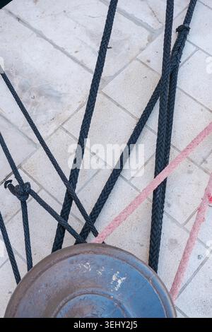 Vue rapprochée de plusieurs épaisses cordes d'amarrage, à la fois foncées et roses, étroitement attachées autour d'une vieille borne rouillée sur un quai pavé. Banque D'Images