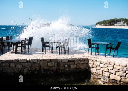 Une forte vague de l'océan éclabousse sur les tables et les chaises de café en plein air sur une jetée de pierre au bord de la mer par une journée claire et claire. Banque D'Images