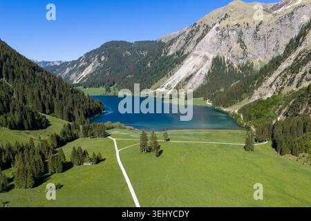 Panorama aérien d'un lac alpin bleu profond encadré par une forêt à feuilles persistantes, des prairies herbeuses et des pentes de montagne abruptes ; des chemins sinueux traversent le premier plan, e. Banque D'Images