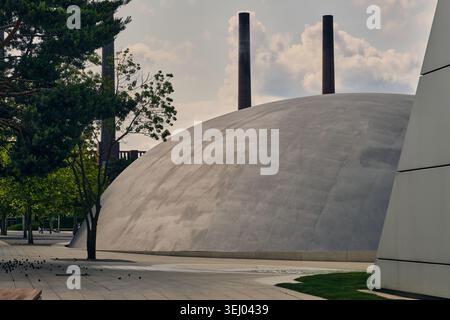 Wolfsburg, Allemagne - 06 juin 2025 : vue du dôme en béton du musée Autostadt avec cheminées industrielles et arbres verts sous un ciel nuageux. Banque D'Images