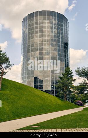 Wolfsburg, Allemagne - 06 juin 2025 : vue de la tour de voiture en verre emblématique d'Autostadt entourée de collines verdoyantes et d'arbres sous un ciel nuageux. Banque D'Images