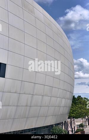 Wolfsburg, Allemagne - 06 juin 2025 : vue de la façade carrelée incurvée du pavillon Autostadt sous un ciel bleu avec nuages et arbres verts. Banque D'Images