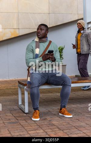 Divers piétons masculins assis sur le banc et passant devant la plaza, tenant des smartphones Banque D'Images