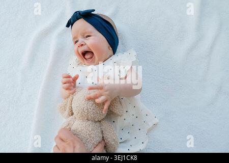Adorable enfant nouveau-né souriant brillamment et jouant avec un jouet lapin en peluche. Belle photo de stock de style de vie d'une fille en bas âge en bonne santé portant un bleu Banque D'Images