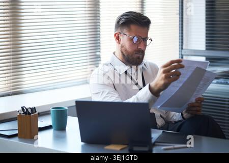 Homme d'affaires caucasien d'âge moyen examinant des documents au bureau, analysant la paperasse à côté de l'ordinateur portable. Idéal pour la finance d'entreprise, l'audit, la conformité, le contenu de gestion Banque D'Images