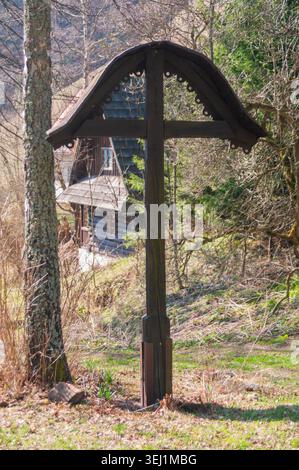 Vue d'une croix traditionnelle en bois avec un toit décoratif devant une cabane historique en rondins dans l'ancien village de Prasnica à Stare Hory, Banskobyst Banque D'Images