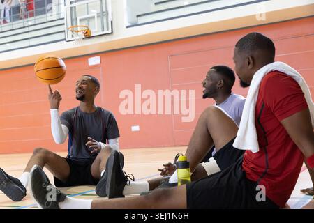 Divers coéquipiers masculins assis sur un terrain de basket-ball, tournoyant basket-ball, portant des vêtements de sport. Groupe, spin, gymnase, camaraderie, détendu, baskets, serviette Banque D'Images