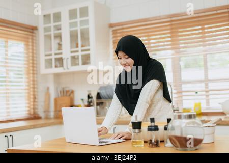 Jeune femme en hijab travaille avec un ordinateur portable à la cuisine à la maison. Elle se concentre sur l'ordinateur portable, tapant calmement. La lumière naturelle remplit l'espace chaleureux de la cuisine en bois. Banque D'Images