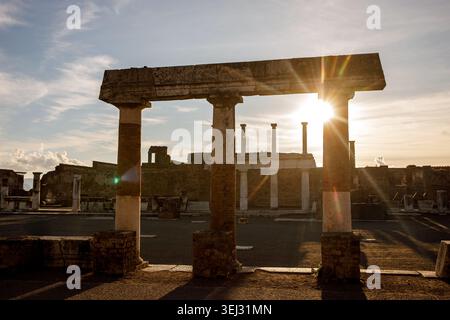 Un portique construit de lourds blocs de pierre se dresse à l'intérieur du site historique de Pompéi, encadré contre un ciel lumineux de coucher de soleil rempli de rayons lumineux. Banque D'Images