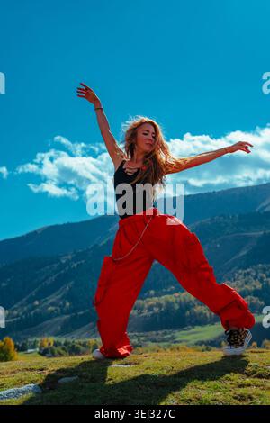Jeune modèle blonde dans une tenue élégante profitant et dansant sur une colline herbeuse avec des montagnes et la forêt derrière Banque D'Images