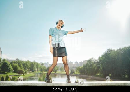 Femme se tient sur le rebord près de la rivière portant chemise bleue et jupe noire. Elle danse librement avec des écouteurs. Les arbres verts et les bâtiments sont visibles à l'arrière Banque D'Images