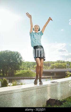 Femme se tient sur le rebord à côté de la rivière avec les bras levés haut. Elle porte une chemise bleue et une jupe noire. Le soleil brille au-dessus. Arbres verts et buildi moderne Banque D'Images
