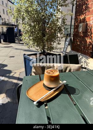 Une boisson au café glacé sur un petit plateau servi sur une table de café verte extérieure avec un arbre vert et une rue ensoleillée de la ville en arrière-plan. Banque D'Images