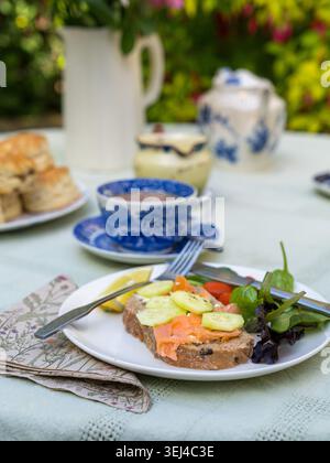 Thé en plein air servi dans un jardin, avec sandwichs ouverts au saumon fumé, feuilles de salade, scones, tasses à thé et théière sur une table sous le soleil d'été. Banque D'Images