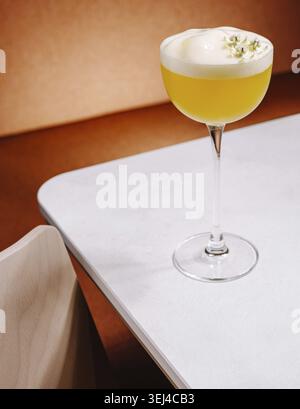 Nourriture, Un cocktail jaune rafraîchissant se trouve élégamment dans un verre à pied sur une table moderne. La boisson comporte un dessus mousseux et des garnitures, invitant les plaisants Banque D'Images