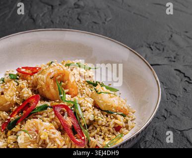 Nourriture, Un bol de riz frit aux crevettes savoureux repose sur une surface sombre. Le plat comprend de succulentes crevettes, des herbes fraîches et des tranches de piment rouge, créant Banque D'Images