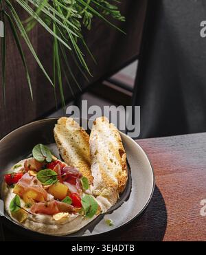 Nourriture, Un bol de houmous garni de légumes colorés et de viande tranchée se trouve à côté de deux morceaux de pain grillé sur une table en bois. La verdure luxuriante ajoute un Banque D'Images