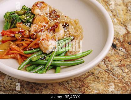 Nourriture, crevettes sautées servies avec des nouilles claires et des légumes vibrants comme le brocoli et les haricots verts. Le plat est présenté élégamment sur un col neutre Banque D'Images