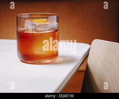Nourriture, Un cocktail est assis sur une table blanche élégante, avec un grand glaçon et une touche d'orange. Le fond chaleureux ajoute une ambiance chaleureuse au settin Banque D'Images