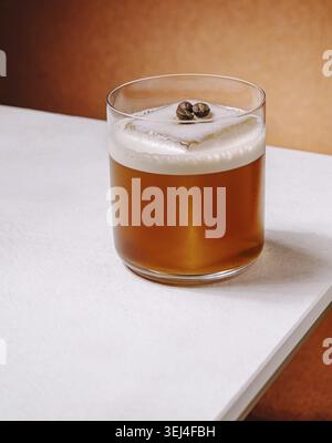 Nourriture, Un cocktail artisanal rafraîchissant se trouve sur une table blanche avec un fond orange chaud. La boisson est recouverte de grains de café et a une couche de mousse crémeuse Banque D'Images
