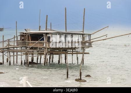 Vieste - Pouilles - Italie - 08-29-2025, une plate-forme de pêche en bois avec cabane et longs poteaux s'étend au-dessus de la mer sous un ciel couvert, avec un shor lointain Banque D'Images