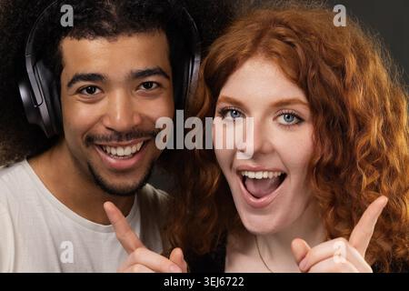 Jeune couple diversifié souriant heureux, posant avec des écouteurs et pointant les doigts Banque D'Images