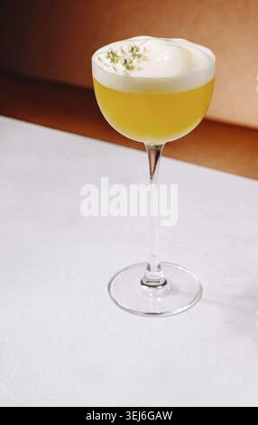 Nourriture, Un cocktail jaune vif se trouve dans un élégant verre, orné d'une garniture mousseuse et garni de petites herbes. La boisson est placée sur un lisse Banque D'Images