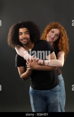 Jeune couple diversifié serrant par derrière, souriant à la caméra sur fond sombre Banque D'Images