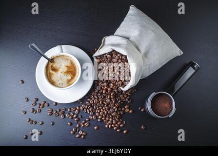 Tasse à café, support d'une machine à expresso et grains de café dans un sac en toile sur fond de table en bois noir. Nature morte avec du café. Vue de dessus Banque D'Images
