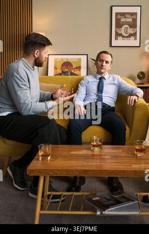 Plan vertical de jeunes hommes adultes assis sur un canapé, discutant de problèmes personnels et écoutant sérieusement, exprimant l'amitié et le soutien masculins. Idéal pour le counseling ou les campagnes de santé mentale pour hommes Banque D'Images