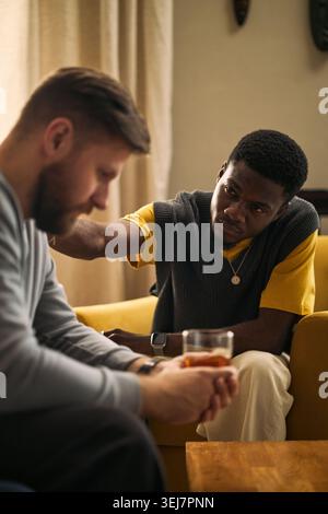 Plan vertical d'un jeune homme noir adulte réconfortant un ami masculin caucasien, offrant un soutien émotionnel pendant une conversation difficile. Utile pour la santé mentale, l'amitié, les campagnes de conseil Banque D'Images