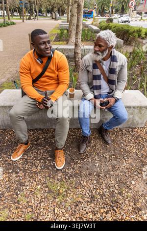 Hommes afro-américains, un aîné, assis sur un banc de béton dans le parc, parlant tout en tenant des téléphones Banque D'Images