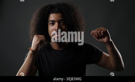 Jeune homme posant avec les poings serrés et expression confiante sur fond sombre Banque D'Images